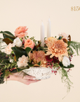 low lying centrepiece with a variety of greenery including peach, white and red flowers, two taper candles. Text reading $150 value