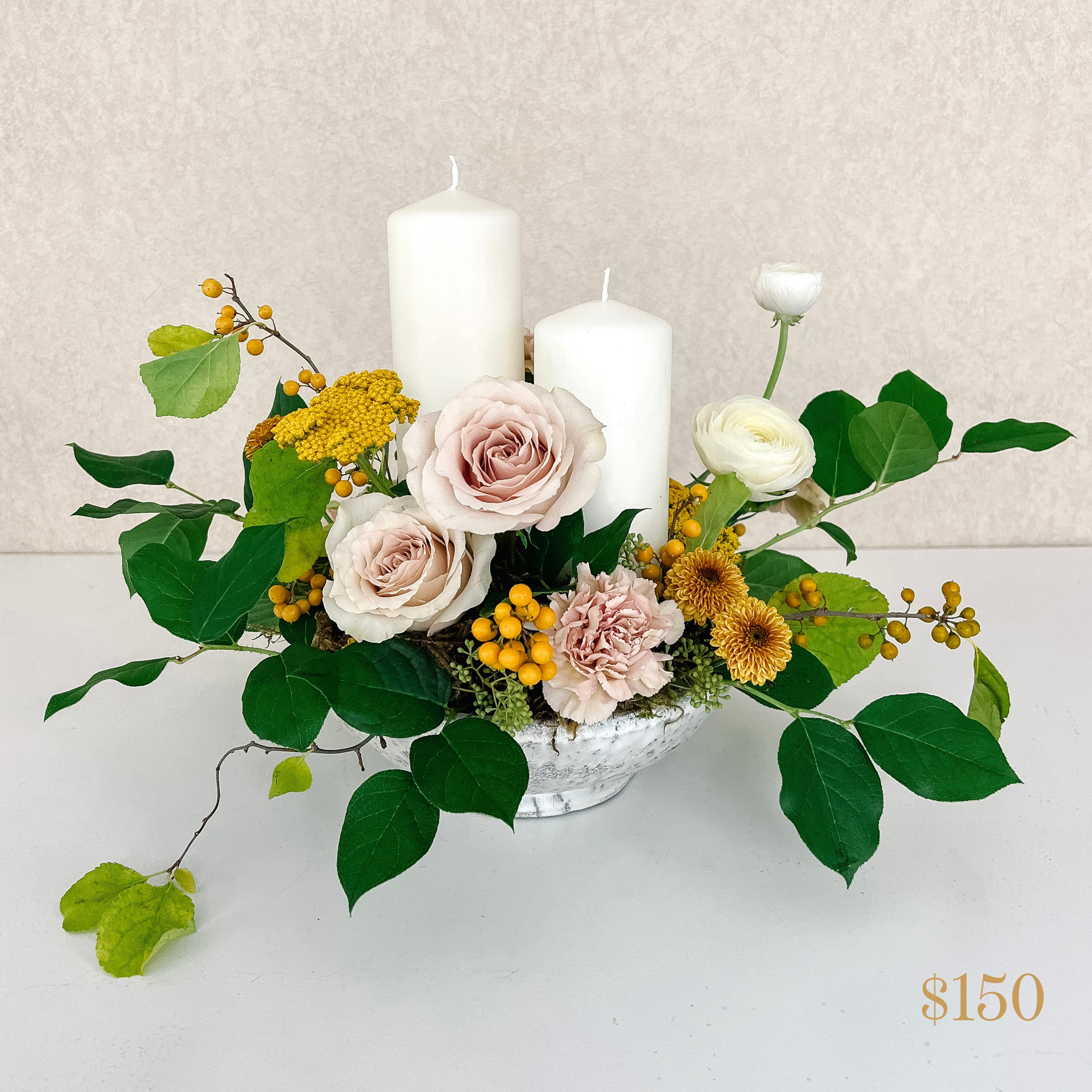 low centrepiece with two pillar candles, a variety of flowers and greenery. Text reading $150 value