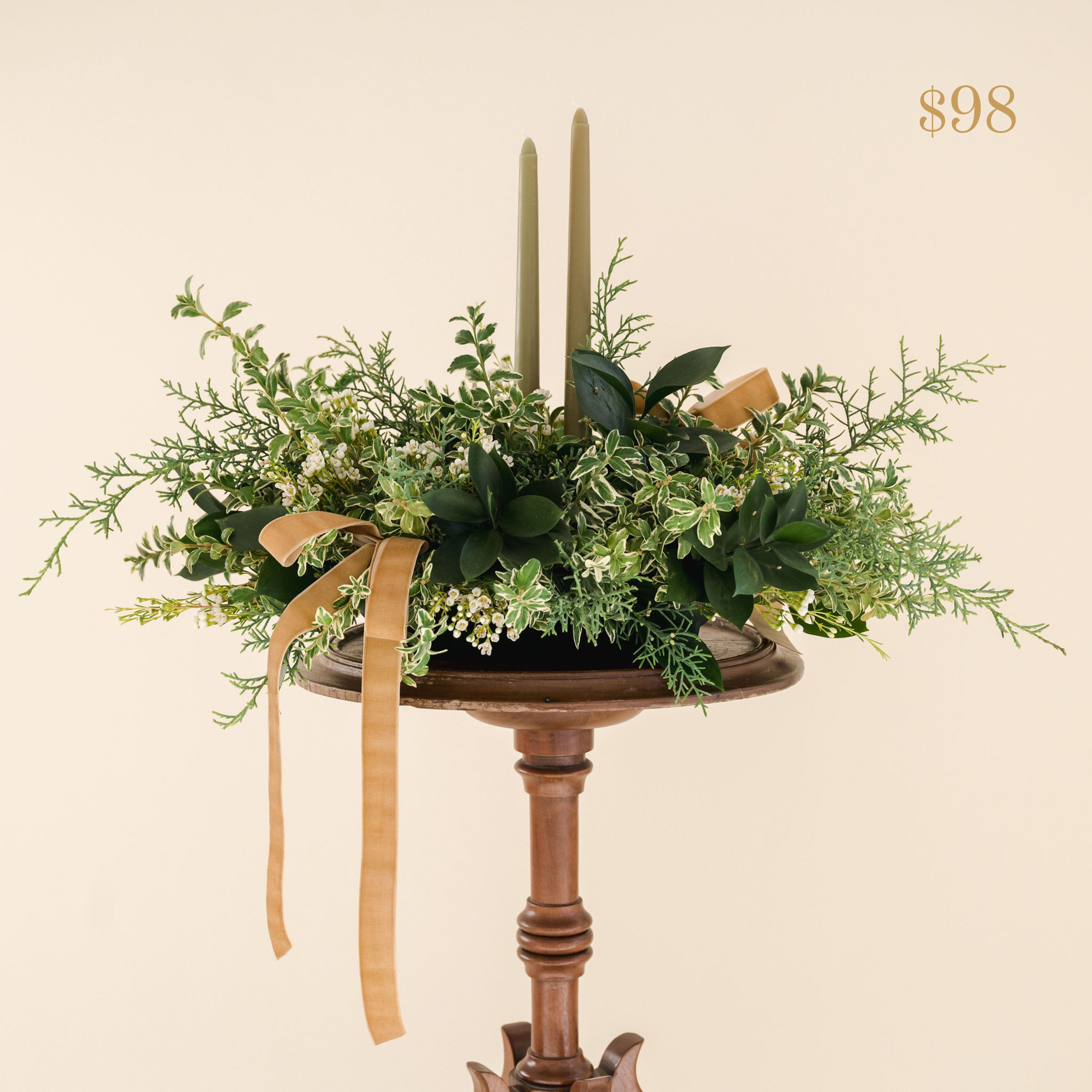 a variety of greenery in a low lying container with two taper candles with ribbon and small white flowers. Text reading $98 value