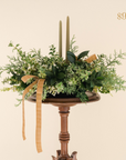 a variety of greenery in a low lying container with two taper candles with ribbon and small white flowers. Text reading $98 value