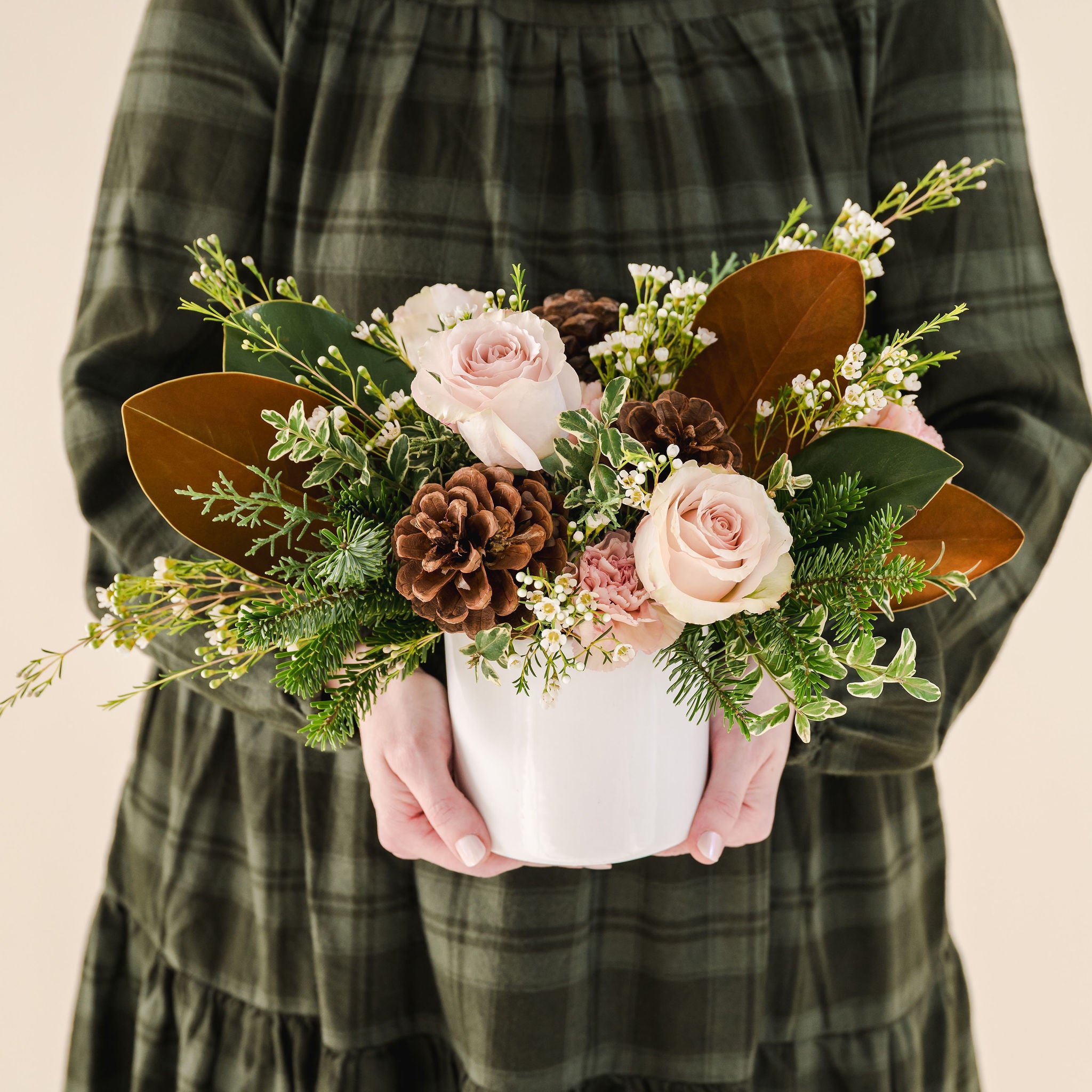 a white ceramic pot with fragrant winter greenery, natural pinecones and beautiful pink, taupe flowers held by a person in a green dress