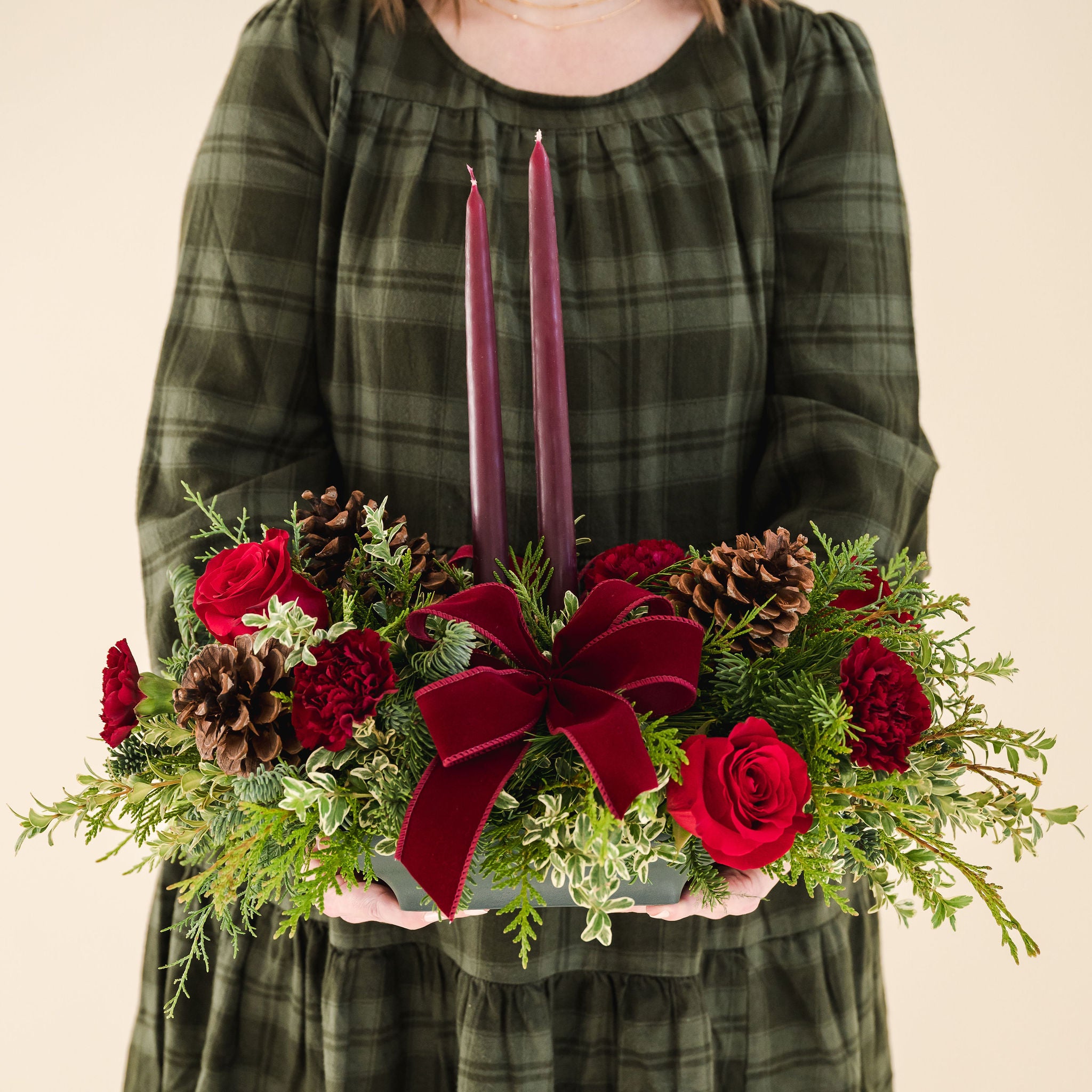 Low arrangement of fragrant winter greens with red and burgundy flowers, two burgundy candles, natural pinecones and burgundy bows being held by a person in a green dress