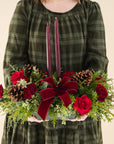 Low arrangement of fragrant winter greens with red and burgundy flowers, two burgundy candles, natural pinecones and burgundy bows being held by a person in a green dress