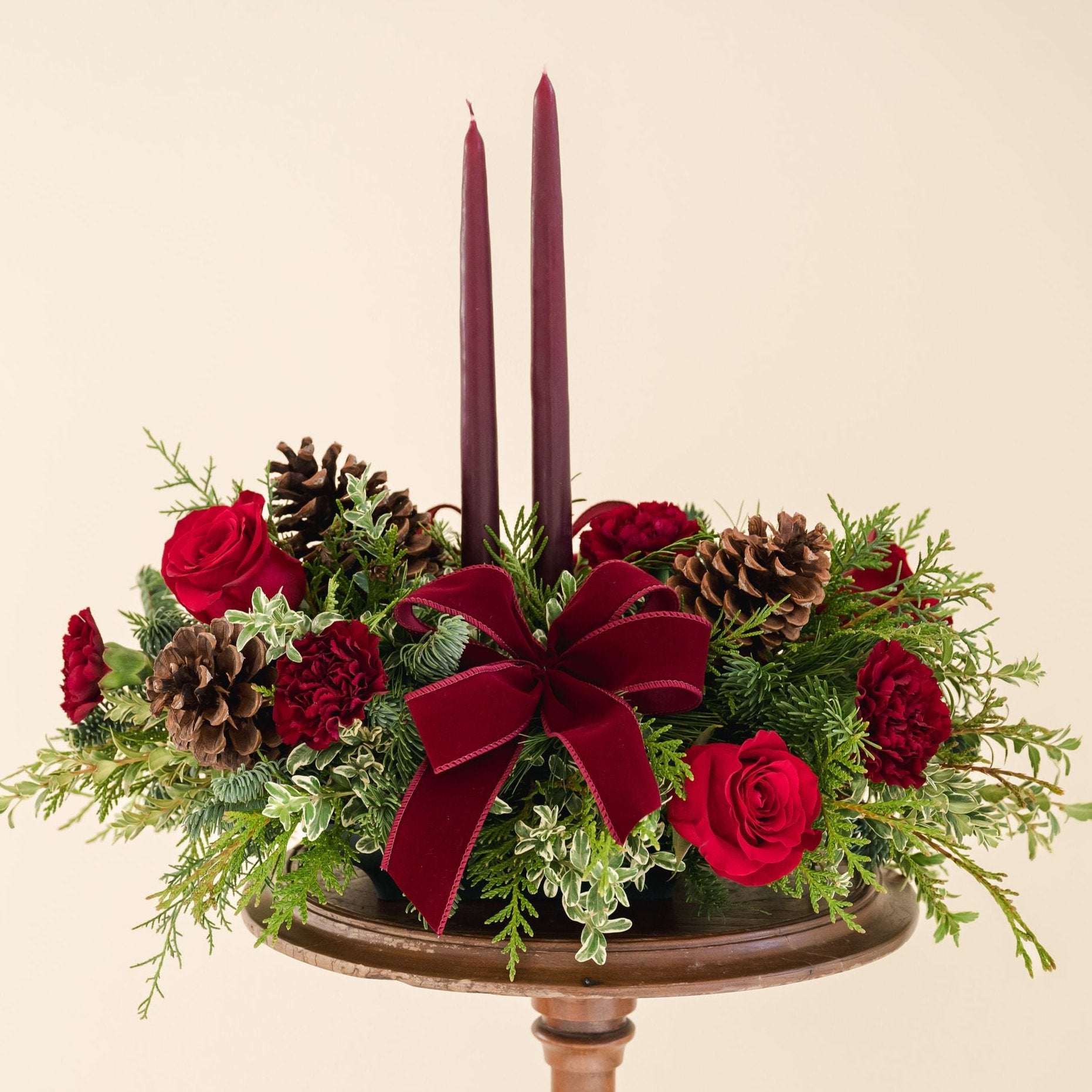 Low arrangement of fragrant winter greens with red and burgundy flowers, two burgundy candles, natural pinecones and burgundy bows.