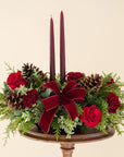 Low arrangement of fragrant winter greens with red and burgundy flowers, two burgundy candles, natural pinecones and burgundy bows.