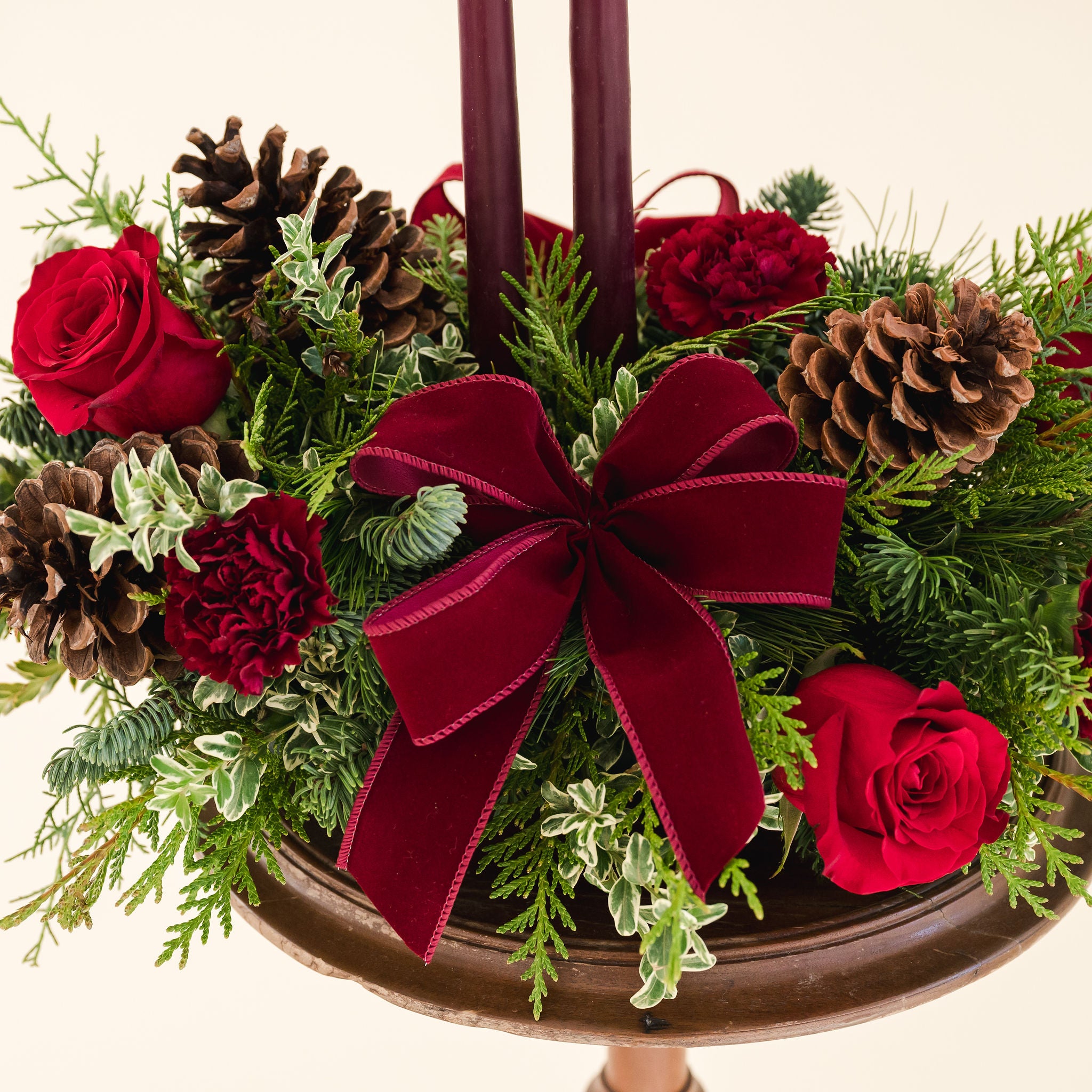 close up of a low arrangement of fragrant winter greens with red and burgundy flowers, two burgundy candles, natural pinecones and burgundy bows.