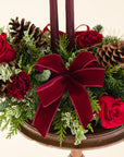 close up of a low arrangement of fragrant winter greens with red and burgundy flowers, two burgundy candles, natural pinecones and burgundy bows.