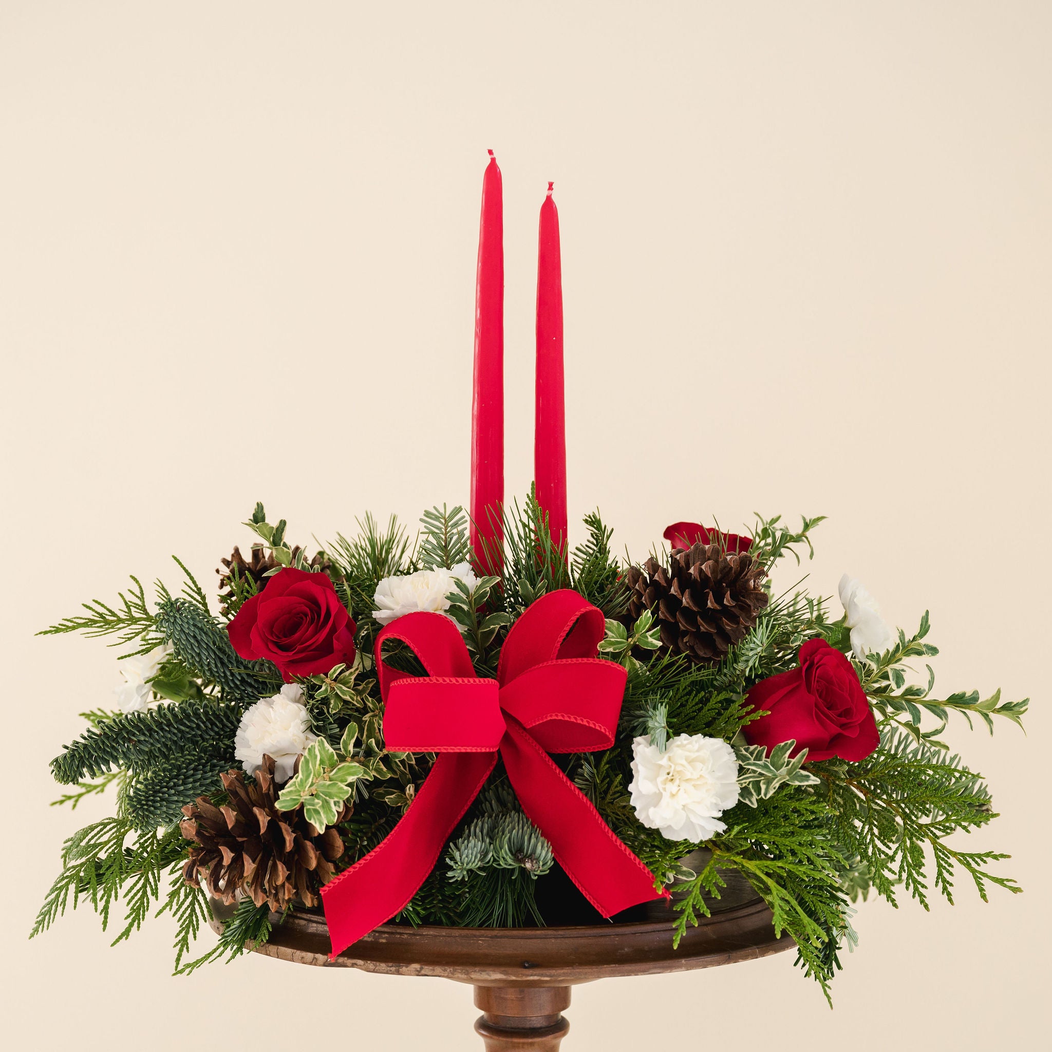 low arrangement of fragrant winter greens with red and white flowers, two red candles, natural pinecones and red ribbon 