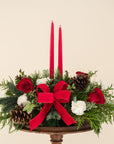 low arrangement of fragrant winter greens with red and white flowers, two red candles, natural pinecones and red ribbon 