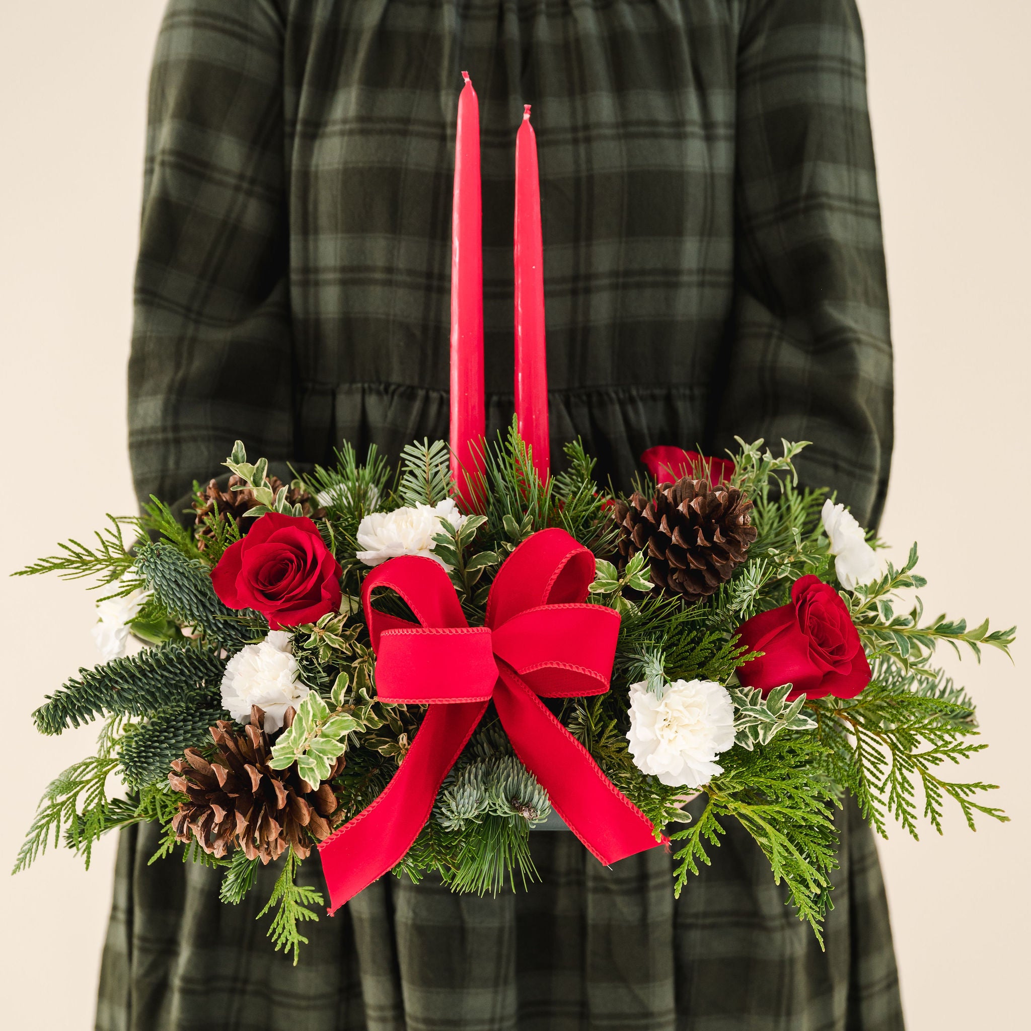 low arrangement of fragrant winter greens with red and white flowers, two red candles, natural pinecones and red ribbon held by person in a green dress