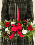 low arrangement of fragrant winter greens with red and white flowers, two red candles, natural pinecones and red ribbon held by person in a green dress