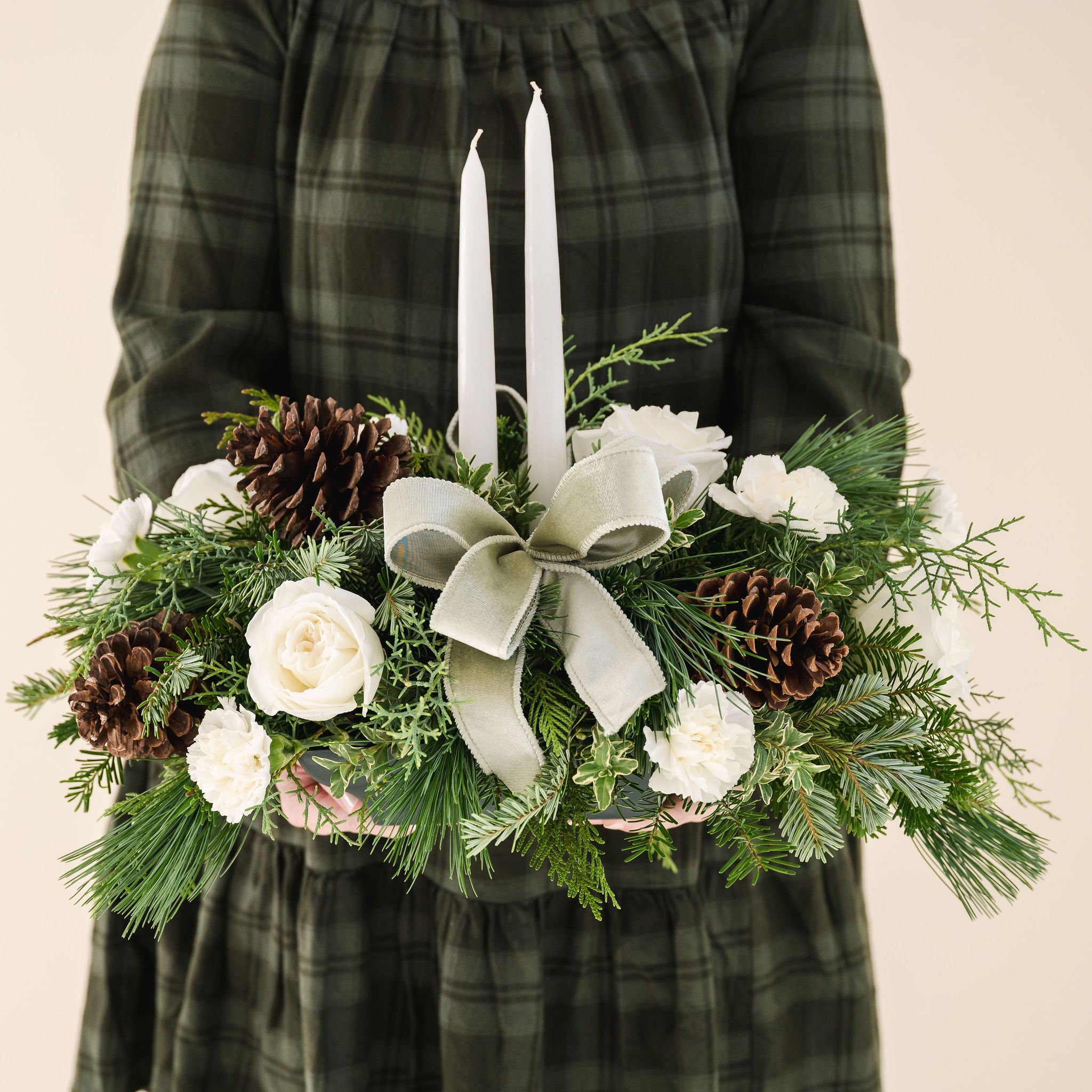 low arrangement of fragrant winter greens with white flowers, two white candles, natural pinecones and sage ribbon held by person in green dress