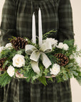 low arrangement of fragrant winter greens with white flowers, two white candles, natural pinecones and sage ribbon held by person in green dress