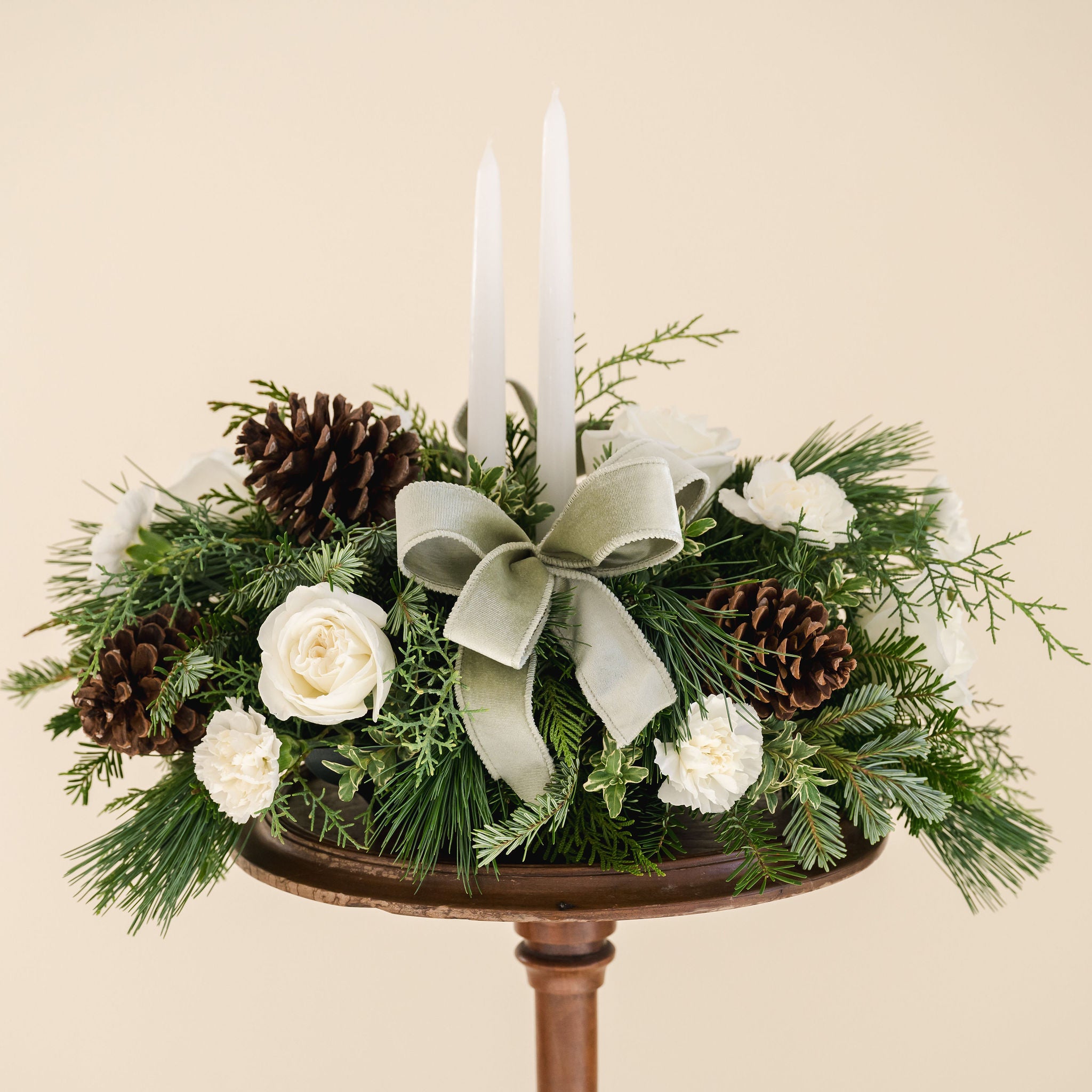 low arrangement of fragrant winter greens with white flowers, two white candles, natural pinecones and sage ribbon. 