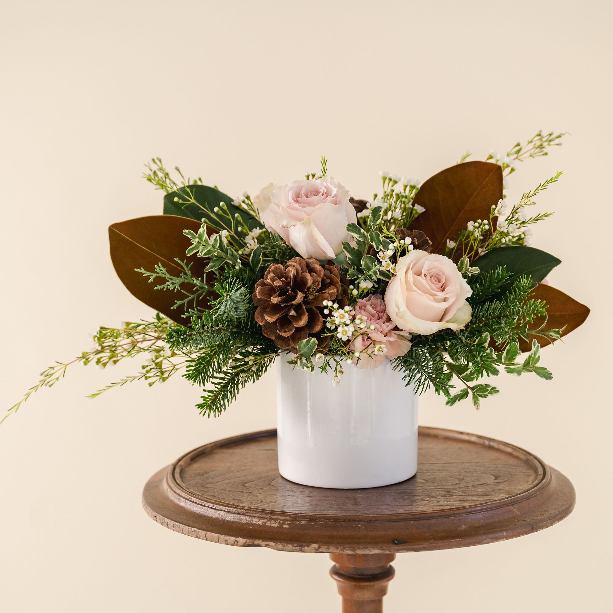 a white ceramic pot with fragrant winter greenery, natural pinecones and beautiful pink, taupe flowers sitting in a wooden stool