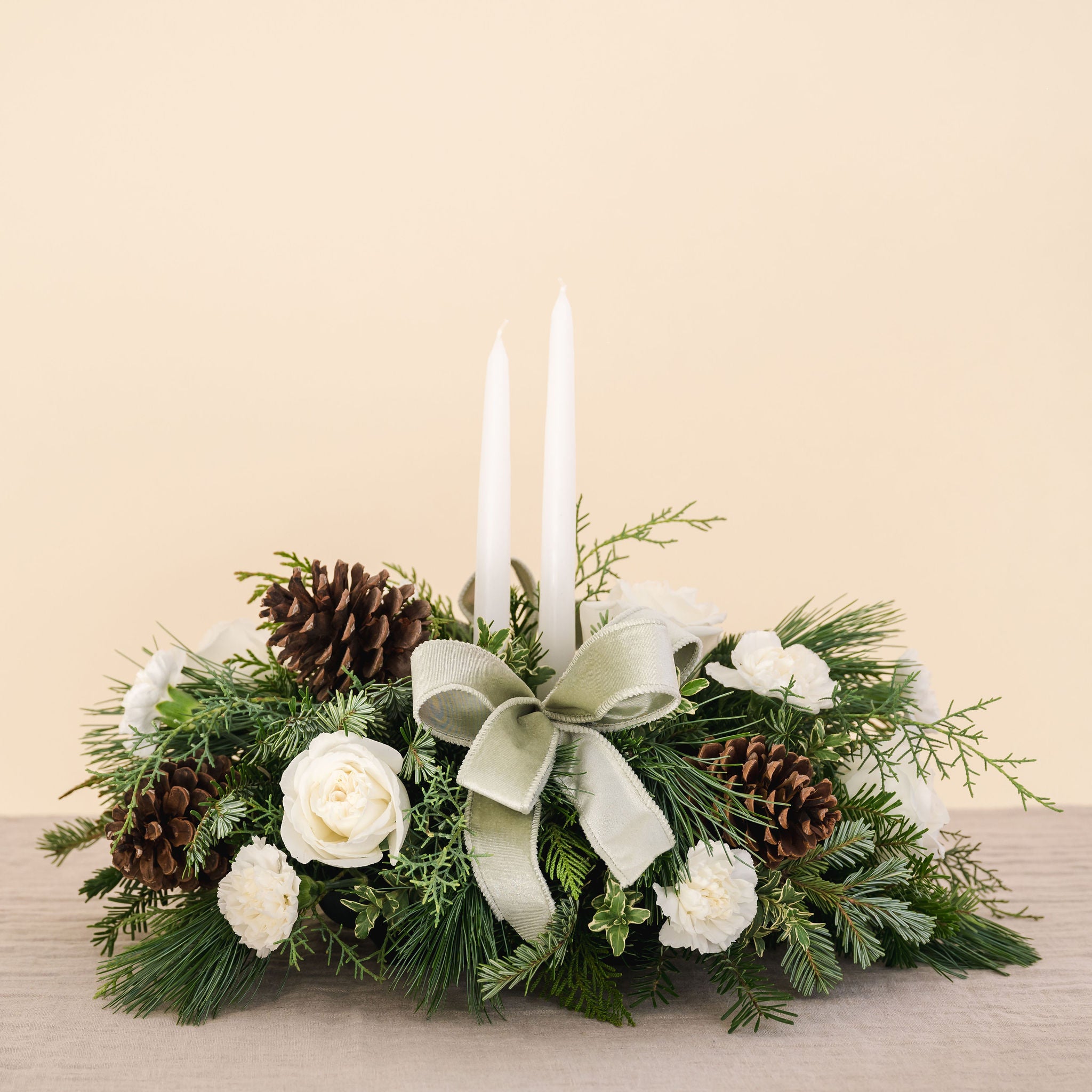 low arrangement of fragrant winter greens with white flowers, two white candles, natural pinecones and sage ribbon.