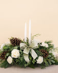 low arrangement of fragrant winter greens with white flowers, two white candles, natural pinecones and sage ribbon.