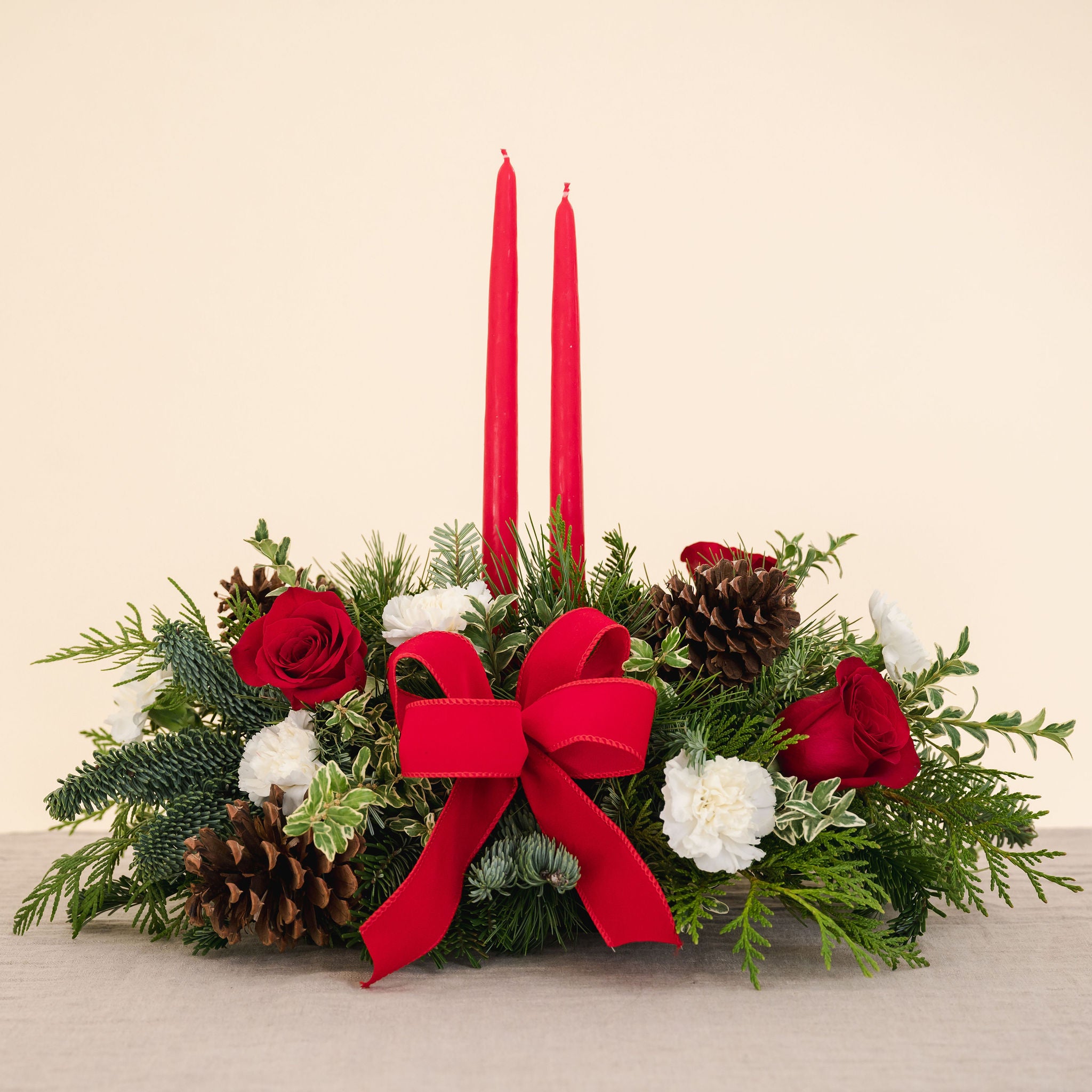 low arrangement of fragrant winter greens with red and white flowers, two red candles, natural pinecones and red ribbon