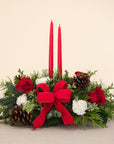 low arrangement of fragrant winter greens with red and white flowers, two red candles, natural pinecones and red ribbon