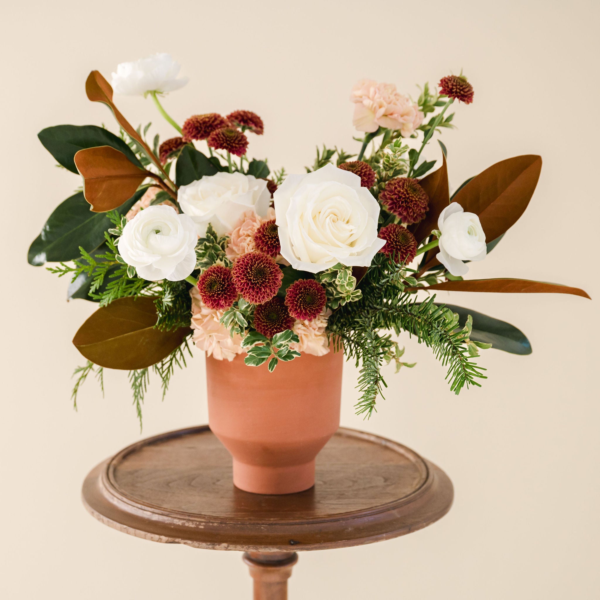 white, rust and peach coloured flowers sit in a bed of greenery, designed in a terracotta vase sitting on a wooden stool