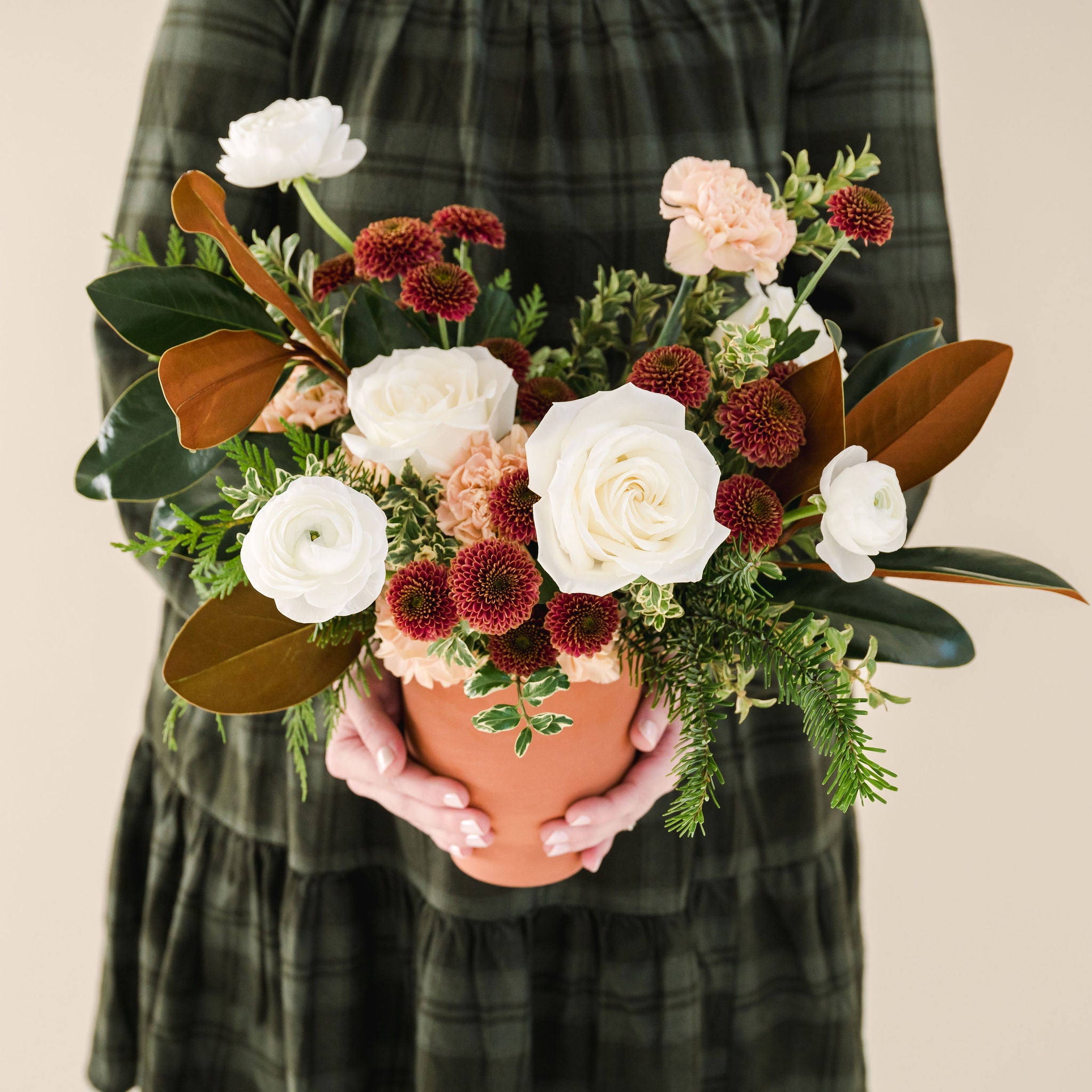 white, rust and peach coloured flowers sit in a bed of greenery, designed in a terracotta vase held by a person in a green dress