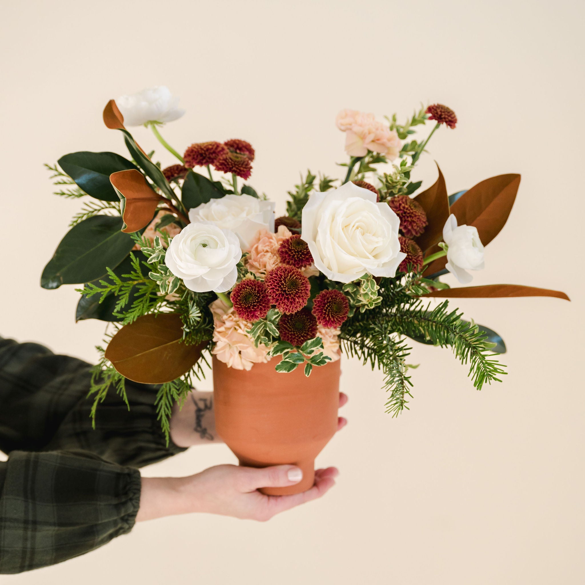 white, rust and peach coloured flowers sit in a bed of greenery, designed in a terracotta vase