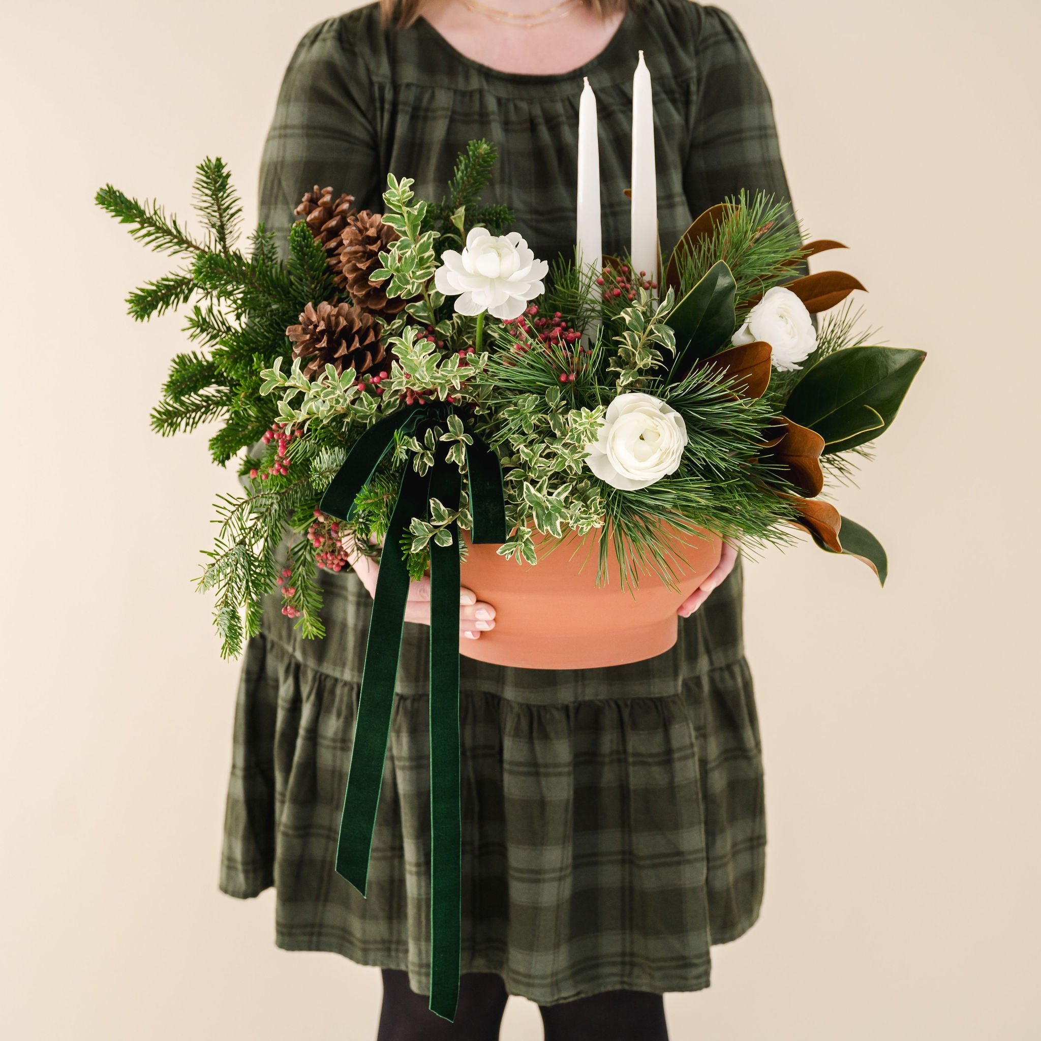 lush, fragrant greens, white ranunculus, 2 white taper candles, natural pinecones and a velvet box designed in a low terracotta vessel, held by a person in a green dress.