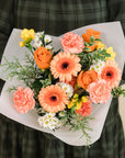 a bouquet of peach, orange, yellow and white flowers with a mix of greenery held by someone in a green dress