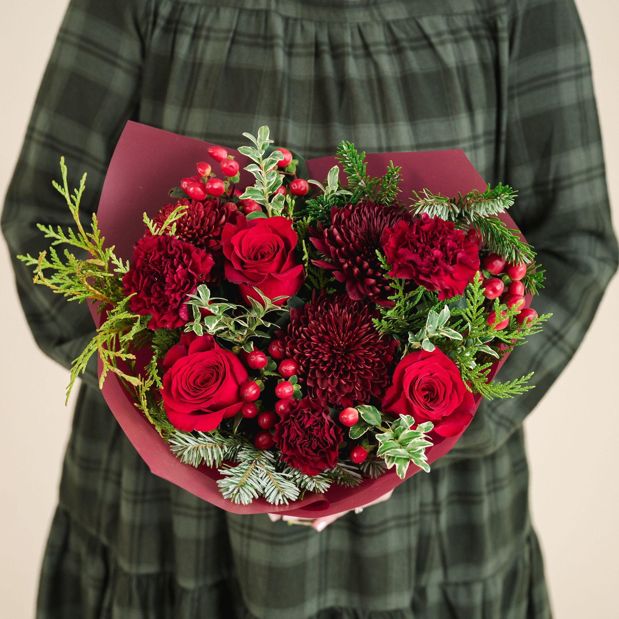 a bouquet of red and burgundy flowers with a mix of fragrant winter greenery, held by someone in a green dress