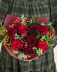 a bouquet of red and burgundy flowers with a mix of fragrant winter greenery, held by someone in a green dress