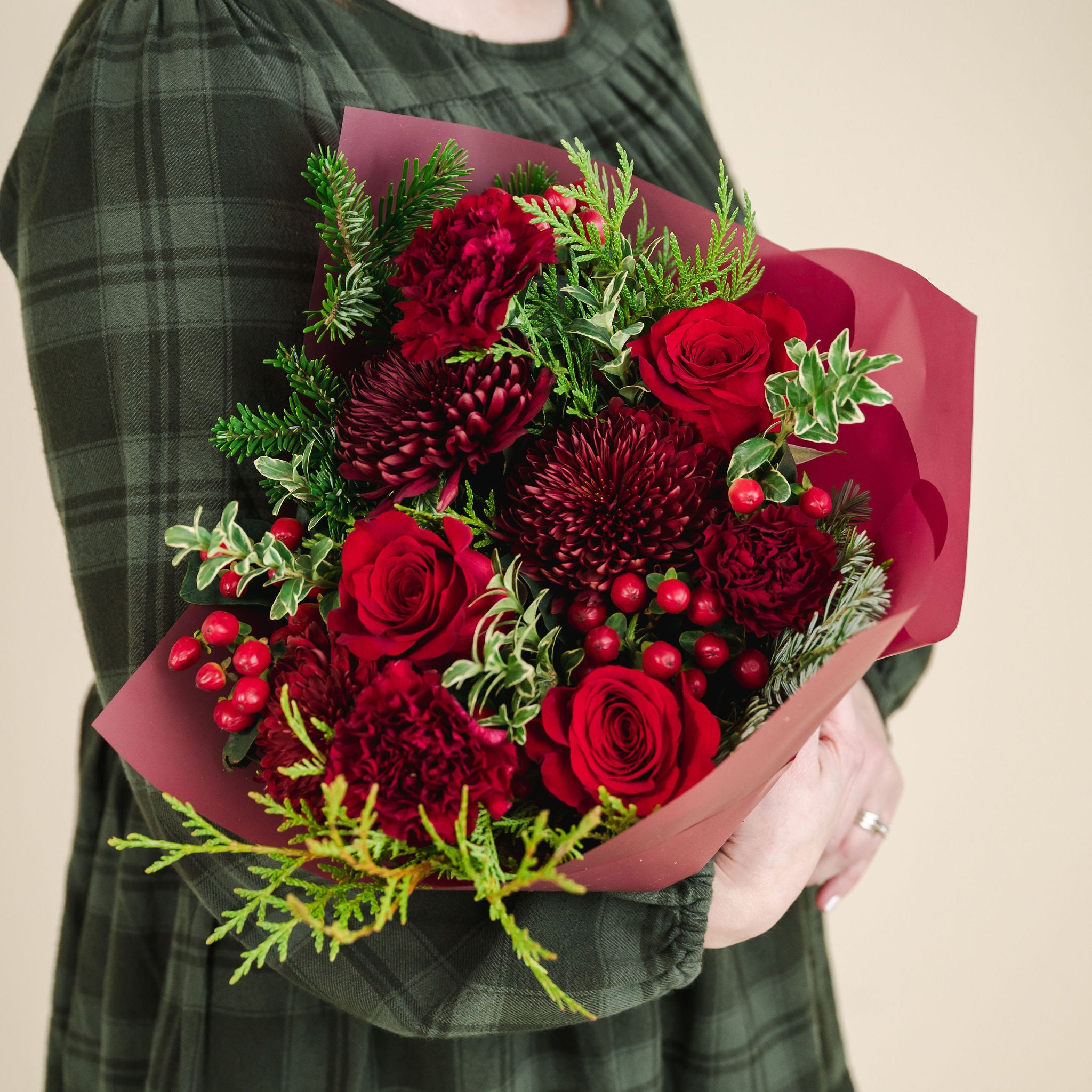 a bouquet of red and burgundy flowers with a mix of fragrant winter greenery, held by someone in a green dress