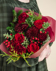 a bouquet of red and burgundy flowers with a mix of fragrant winter greenery, held by someone in a green dress