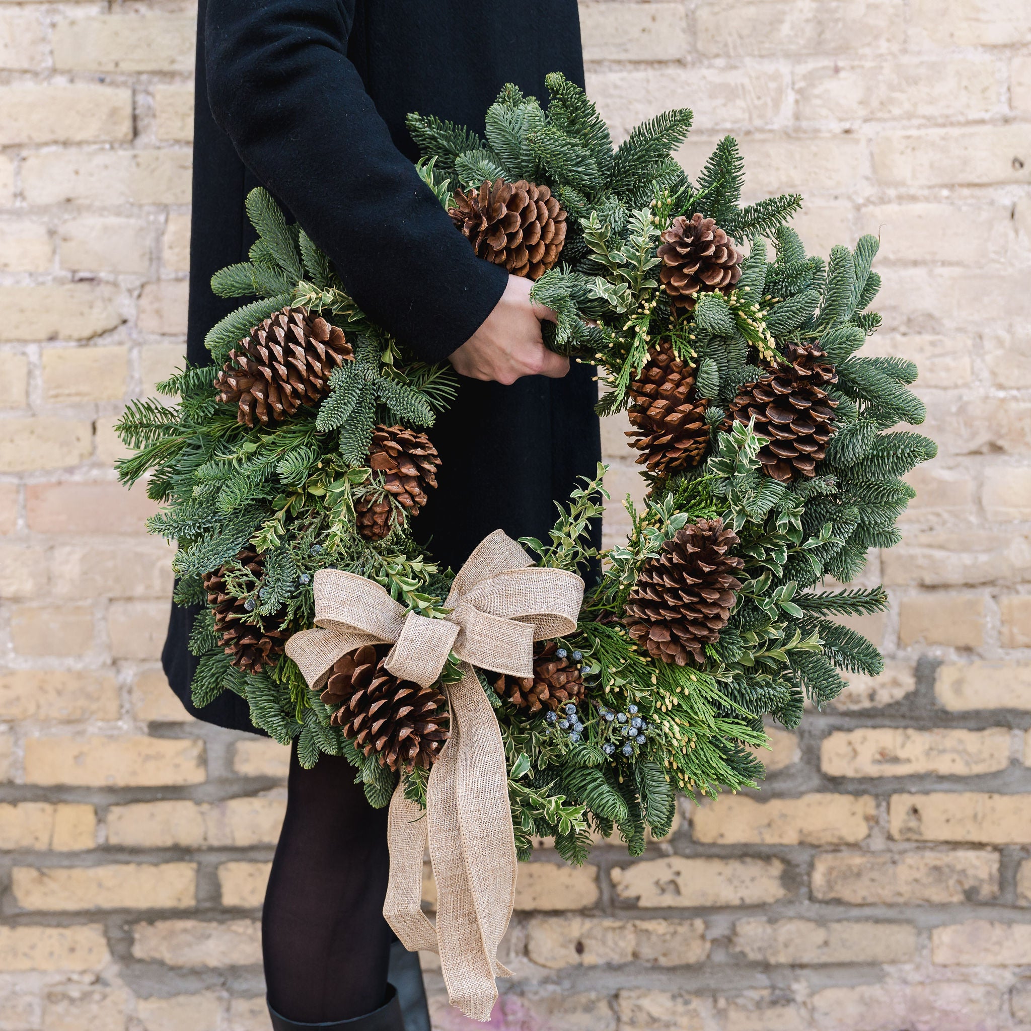 a wreath made of fragrant winter greens, natural pinecones and a burlap bow held by a person in a black coat