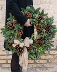 a wreath made of fragrant winter greens, natural pinecones and a burlap bow held by a person in a black coat