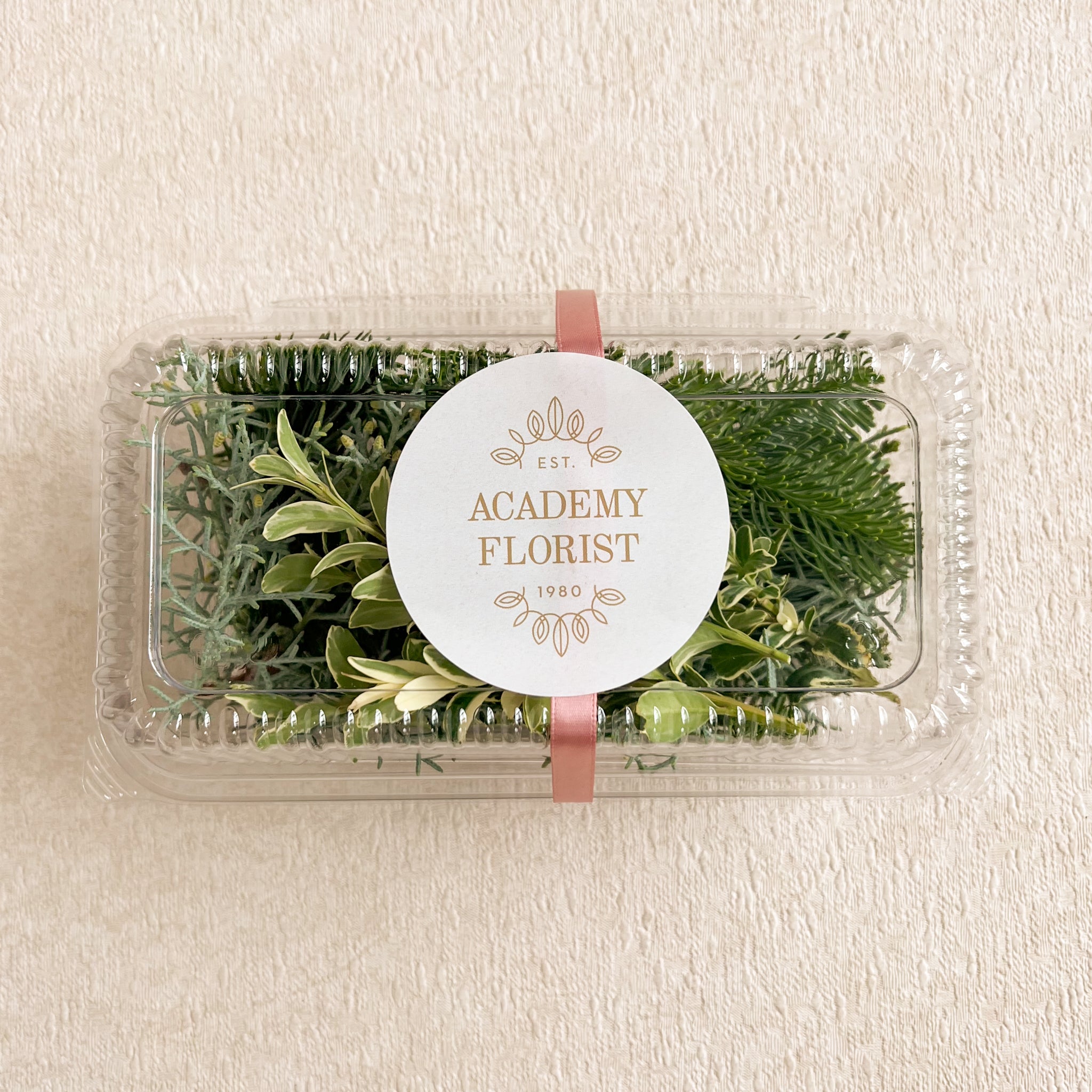 a clear box with mixed winter greenery bits, wrapped with a peach ribbon and secured with an Academy Florist sticker